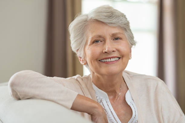 Smiling older woman with short gray hair sitting comfortably on a couch, wearing a light cardigan and a necklace.
